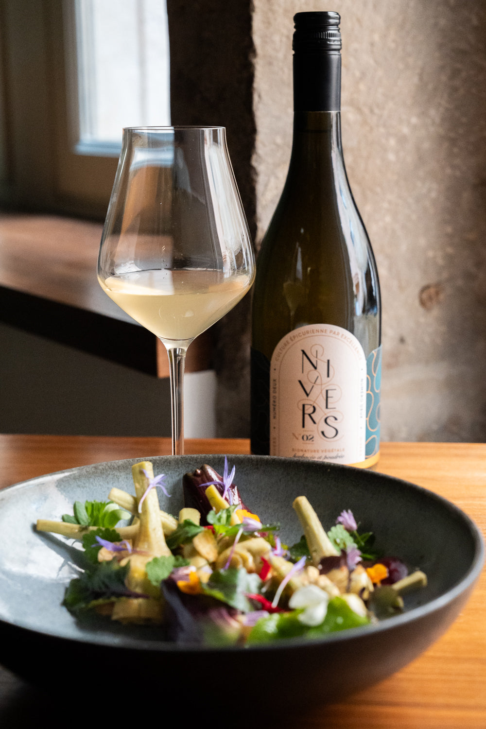 A bottle of Nivers 02 non -alcoholic cuvée of Grand cru Green Tea from Oita Japan, French Verjus, Kombucha culture stands next to a wine glass and a dish of salad on a wooden table.