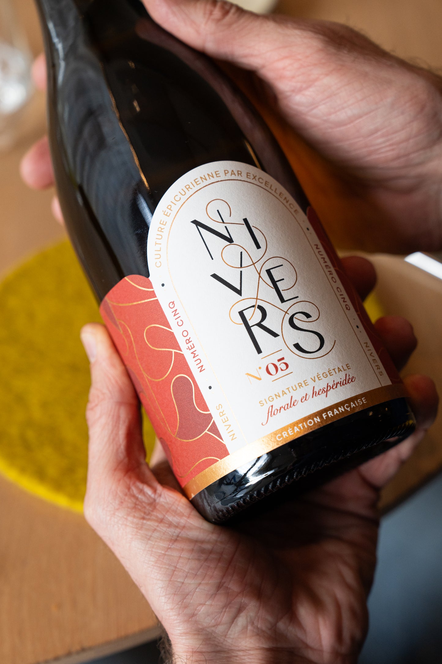 A person holds a  bottle of Nivers No. 05 non-alcoholic cuvée of Grand cru of Fujian and Chinese white and jasmine green teas with both hands against a warm, softly blurred background.