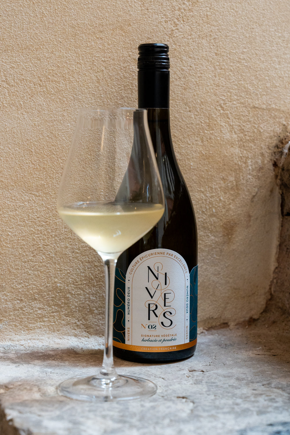 A bottle of Nivers 02 non -alcoholic cuvée of Grand cru Green Tea from Oita Japan, French Verjus, Kombucha culture stands next to a filled wine glass against a rustic beige plaster wall.