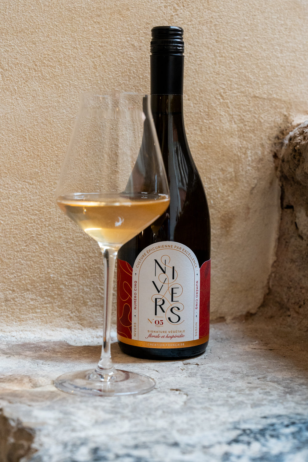 A bottle of Nivers No. 05 non-alcoholic cuvée of Grand cru of Fujian and Chinese white and jasmine green teas next to a filled wine glass on a stone surface against a plastered beige wall.