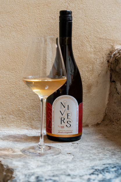 A bottle of Nivers No. 05 non-alcoholic cuvée of Grand cru of Fujian and Chinese white and jasmine green teas next to a filled wine glass on a stone surface against a plastered beige wall.