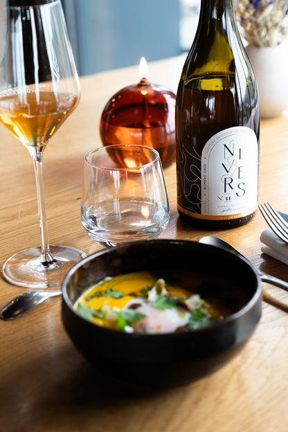 A bottle of Nivers N11 sophisticated non-alcoholic cuvée of Grand Cru black tea, Japanese green tea, French Verjus, and Osmanthus flower  stands on a wooden table beside a wine glass, an empty tumbler, a bowl of soup, and a lit amber candle.