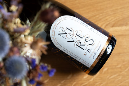 A bottle of Nivers N11 sophisticated non-alcoholic cuvée of Grand Cru black tea, Japanese green tea, French Verjus, and Osmanthus flower lies diagonally on a light wood surface next to blurred dried flowers. The bottle has a dark glass body with a white oval label featuring elegant black typography.